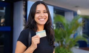 Uma pessoa sorridente comemora a conquista da CNH, segurando o documento com orgulho em um ambiente externo e institucional